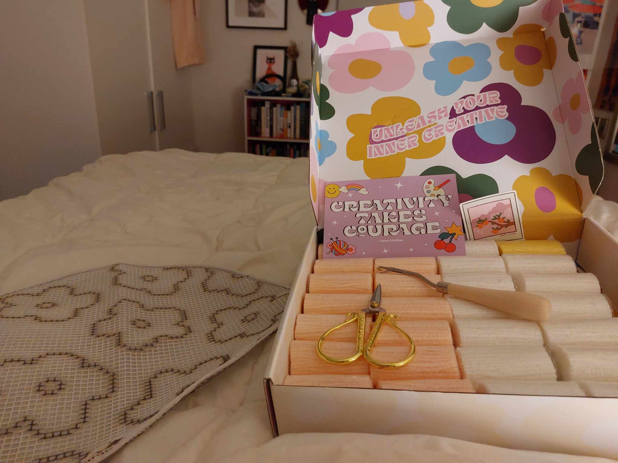 An opened rug making kit, displayed on a bed. It contains lots of little bundles of yarn, a latch hook, a gold pair of scissors and a postcard saying "creativity takes courage". The box is decorated with an abstract flower design and says "unleash your inner creative". The latch hook canvas sits on the bed next to the open kit. 