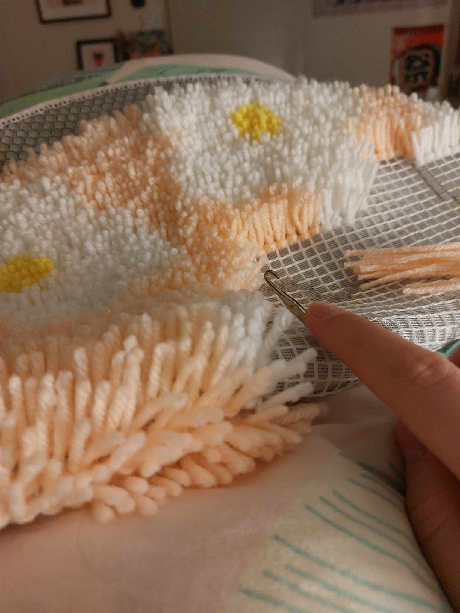 Rug making in progress. The rug canvas is folded over and about half completed with lots of little yarn nots. A hand displays the latch hooking tool next to it. 