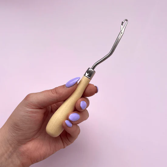 A hand with purple nail polish holds a latch hook against a pink background. 